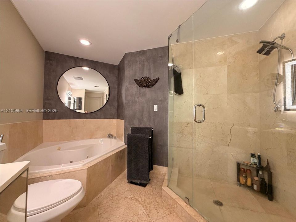 Jetted corner soaking tub, walk-in glass-enclosed shower with stone-finish tiling, and circular wall-mounted mirror