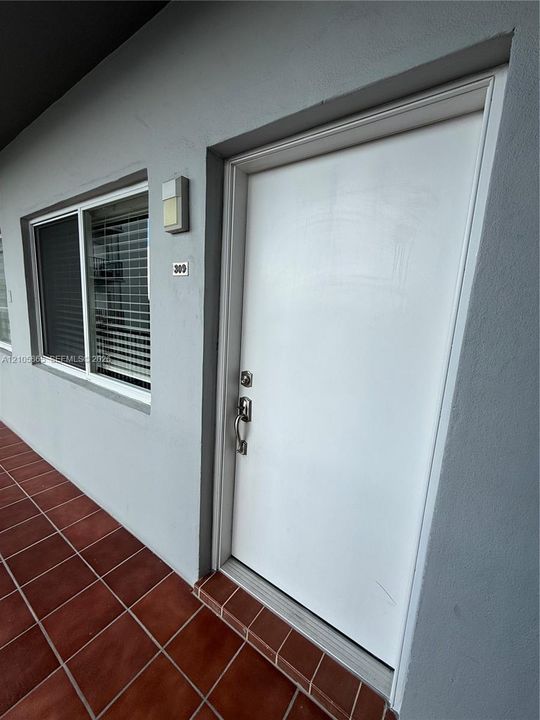 Entryway featuring a solid white door with a silver-finish handle set, terracotta-toned tile flooring, and exterior wall-mounted lighting