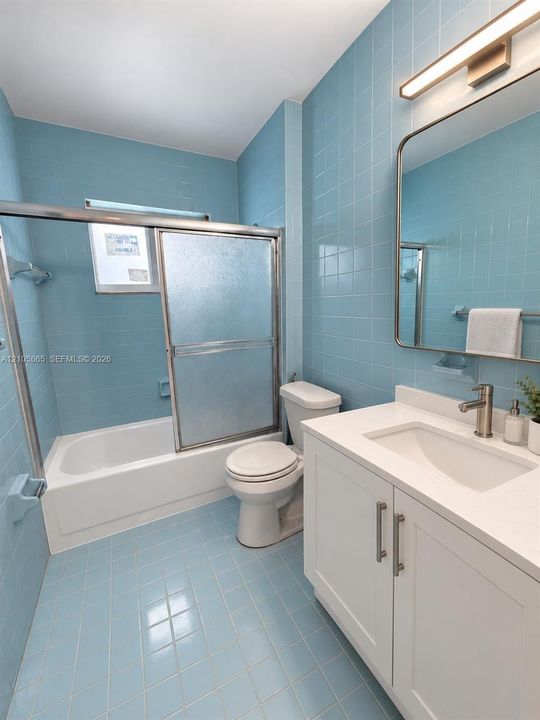 Bathroom featuring light blue wall and floor tiling, white vanity cabinet with a stone countertop, under-mount sink, integrated bathtub and shower combination with sliding glass doors, and a rectangular wall-mounted mirror with integrated lighting