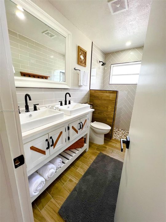 Main House Double vanity featuring vessel sinks, matte black hardware, wood-finish flooring, herringbone-patterned wall tile, and a walk-in shower