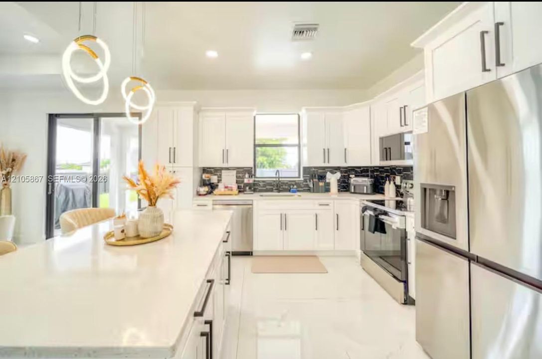 Kitchen featuring white shaker-style cabinetry, a quartz waterfall island, stainless steel appliances, a dark tile backsplash, and contemporary sculptural pendant lighting