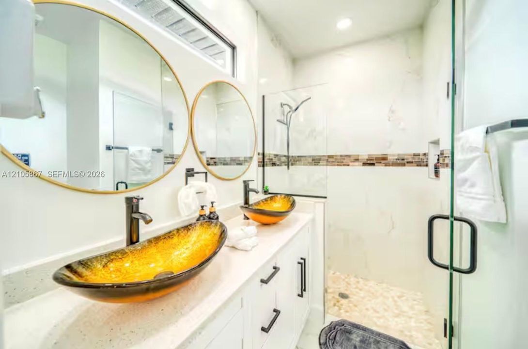 Dual vanity featuring white cabinetry, light-toned stone-finish countertops, vessel sinks, round mirrors with metallic frames, and a walk-in shower with stone tile flooring