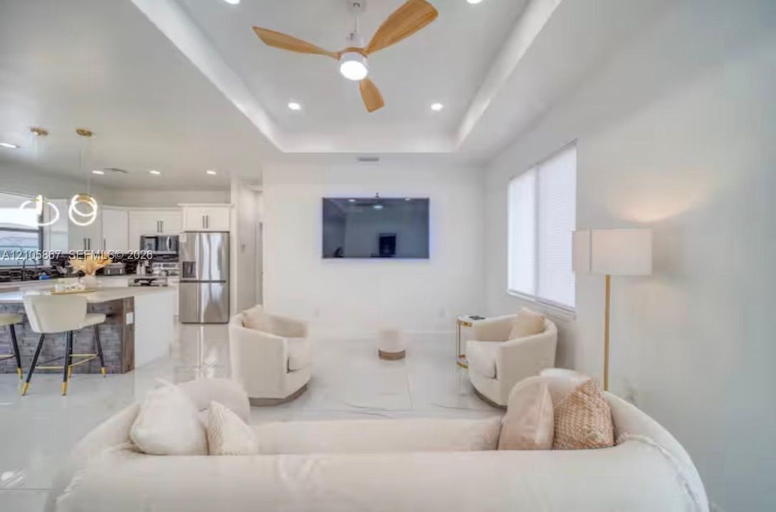 Open-concept interior featuring a recessed tray ceiling, three-blade wood-finish ceiling fan, recessed lighting, white tile flooring, and a kitchen island with stone-finish base