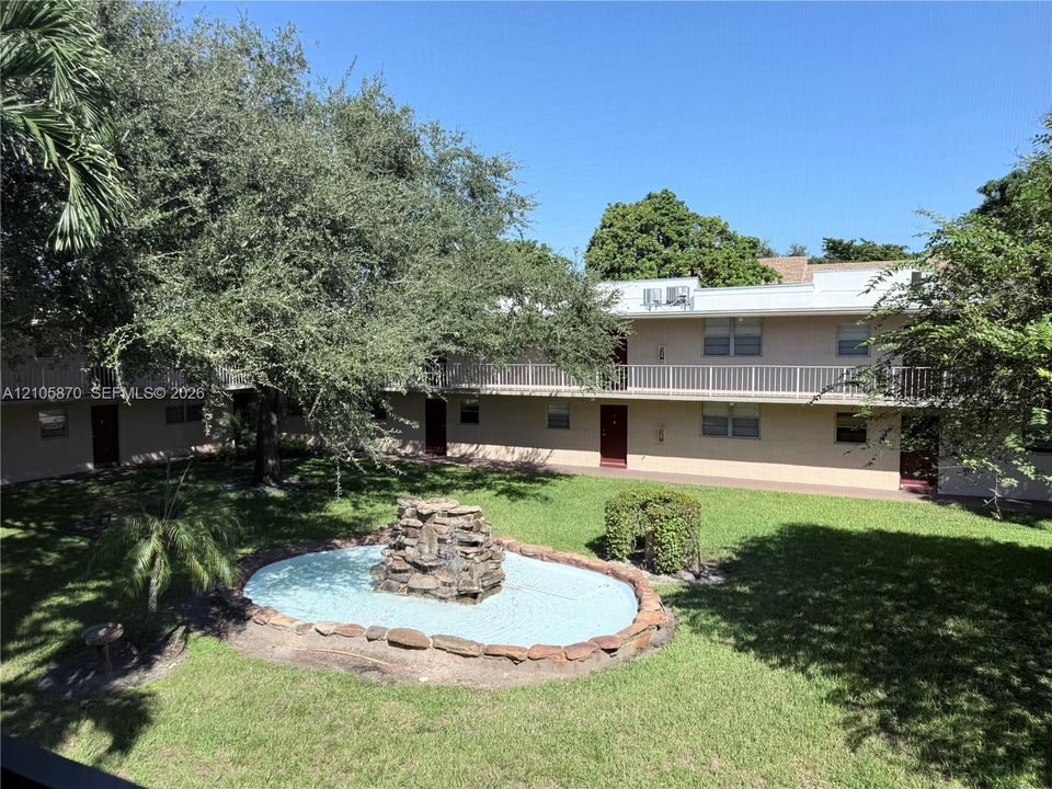 Courtyard featuring a stone-accented fountain, manicured lawn, mature trees, and multi-level exterior walkways with railings