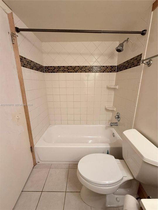 Full bathroom featuring a tub-shower combination with square white wall tiling, a decorative mosaic tile accent border, integrated soap shelving, and tiled flooring