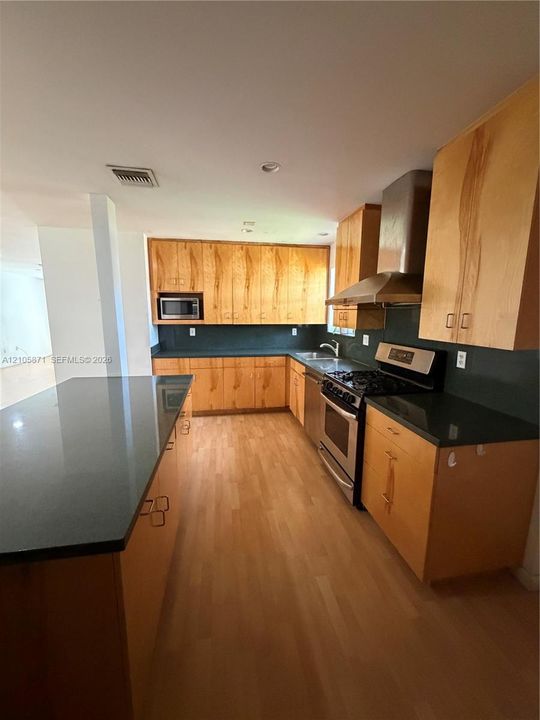 Kitchen featuring light wood-finish cabinetry, dark solid-surface countertops, wood-finish flooring, stainless steel range with matching vent hood, and a built-in microwave