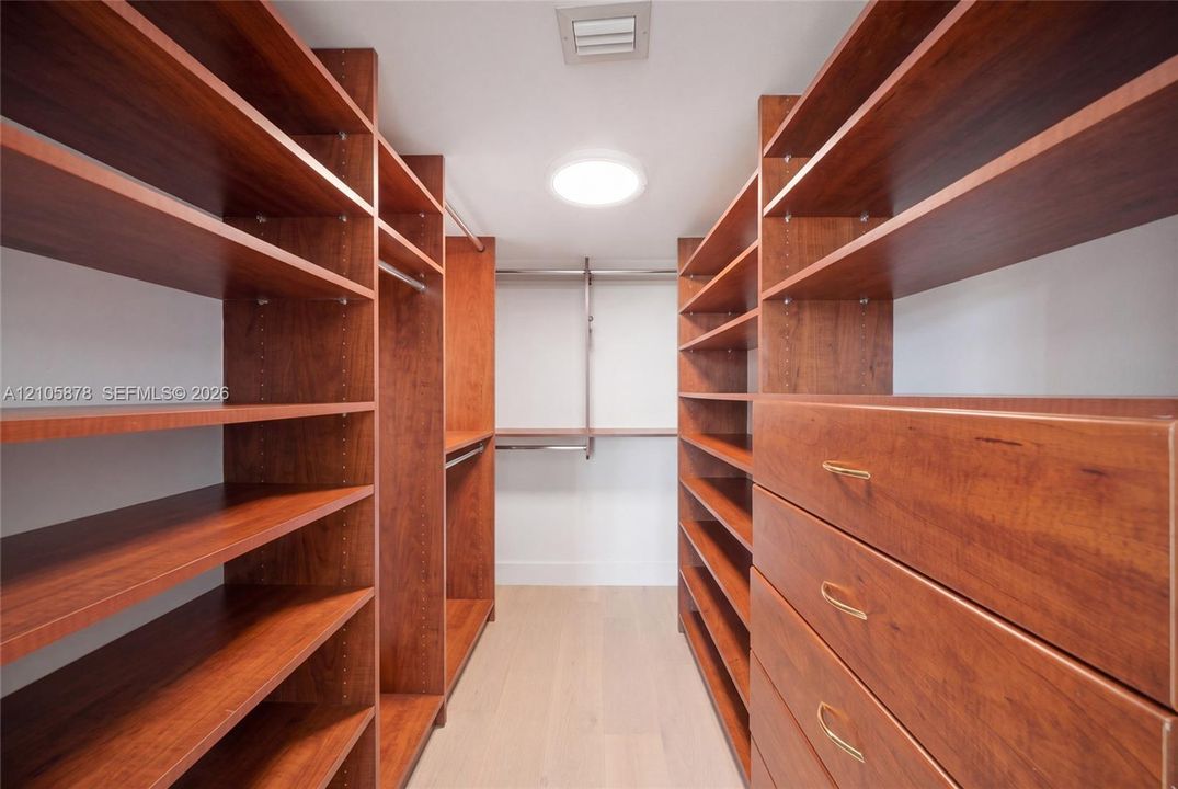 Walk-in storage space featuring extensive custom wood-finish shelving, integrated clothing rods, built-in dresser drawers with metallic pulls, and light-toned flooring