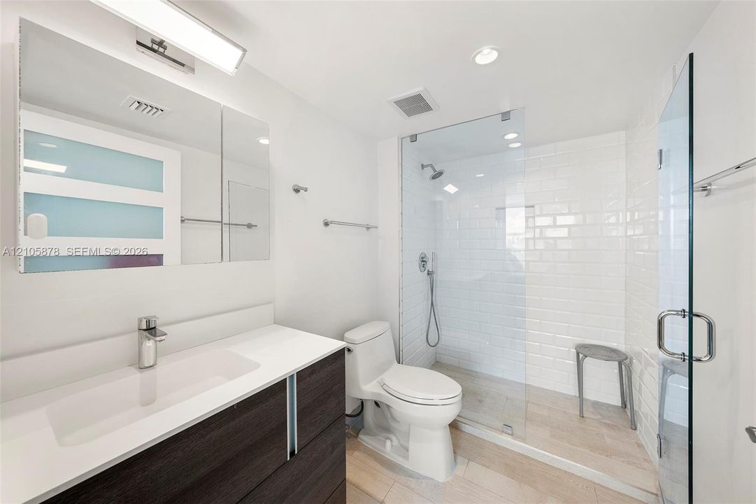 Dark-finish floating vanity with a white countertop and rectangular integrated sink, walk-in shower with white subway tile surround and glass enclosure, wall-mounted medicine cabinet with integrated lighting, and light-toned tile flooring