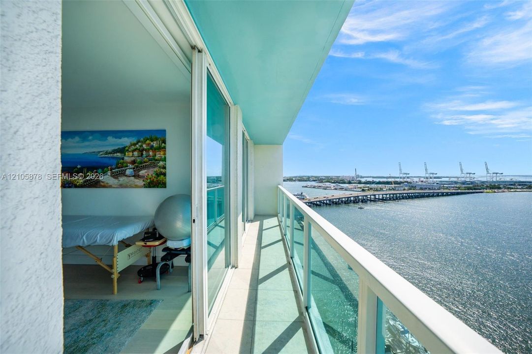 Covered balcony with a glass panel railing, featuring large sliding glass doors and neutral tile flooring