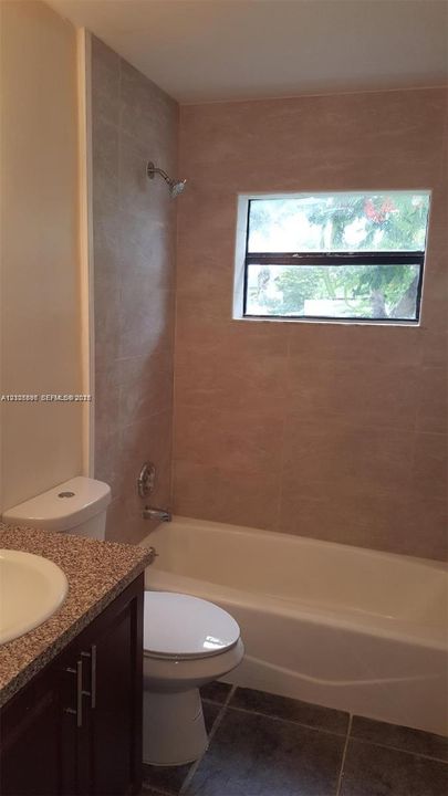 Bathroom featuring a dark-finish vanity, stone-patterned countertop with integrated sink, porcelain tile tub surround, and dark-toned floor tile