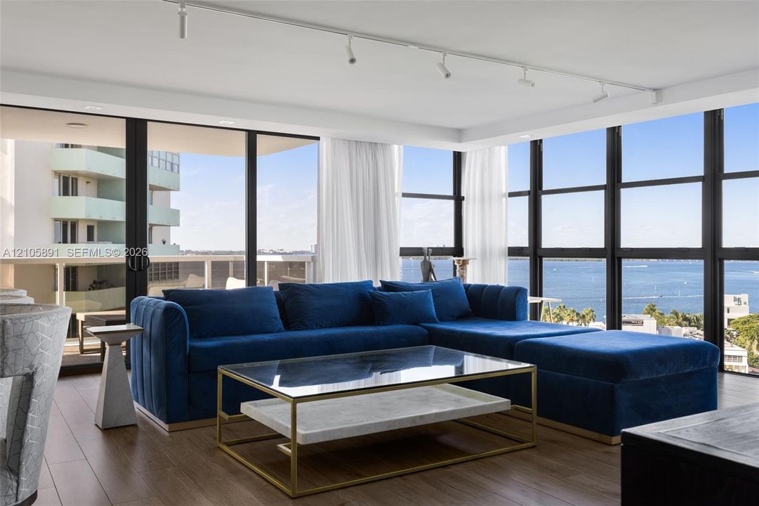 Floor-to-ceiling windows with black metal frames, wood-finish flooring, ceiling-mounted track lighting, and a sliding glass door opening to a private balcony