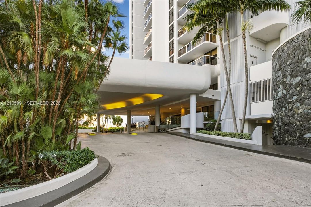 Covered porte-cochère entry featuring stone masonry accents, a concrete drive, and mature palm landscaping