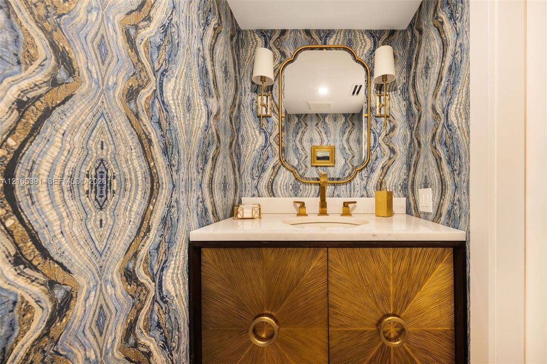 Custom vanity with metallic-textured cabinetry, stone countertop, integrated undermount sink, brass-finish fixtures, and designer wallpaper