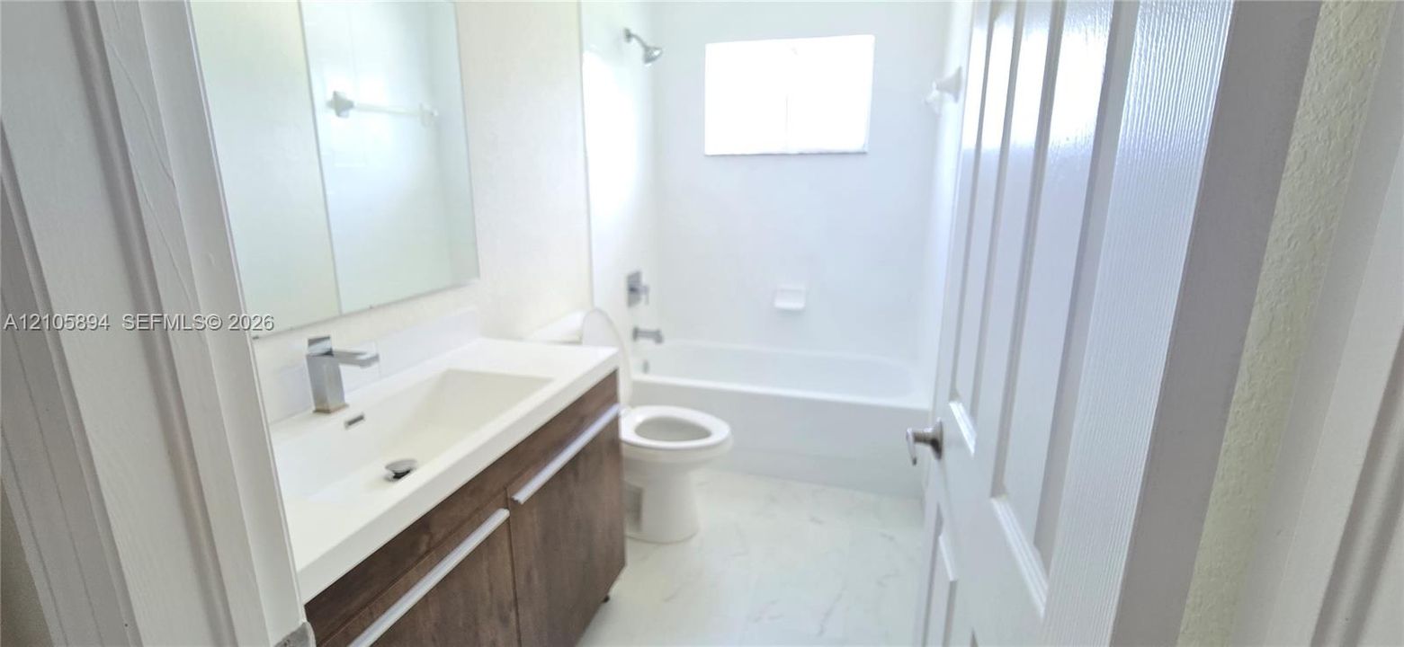 Bathroom featuring a vanity with wood-finish cabinetry, a white integrated basin, a single-handle chrome faucet, a rectangular wall-mounted mirror, and a porcelain-surround tub with a shower head