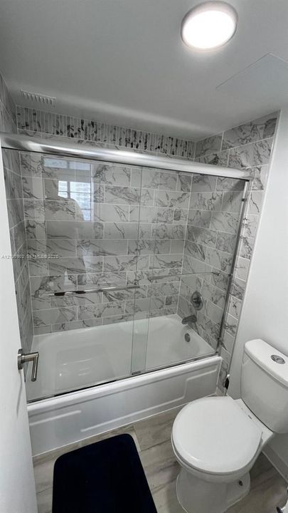 Bathroom featuring a soaking tub with a glass sliding enclosure, floor-to-ceiling stone-finish tile surround, wood-finish flooring, and a flush-mount ceiling light fixture