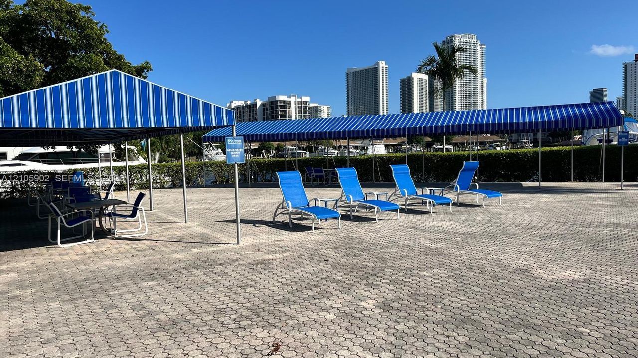 Outdoor paved patio area featuring extensive striped retractable awnings, perimeter privacy hedges, and paved stone surfacing