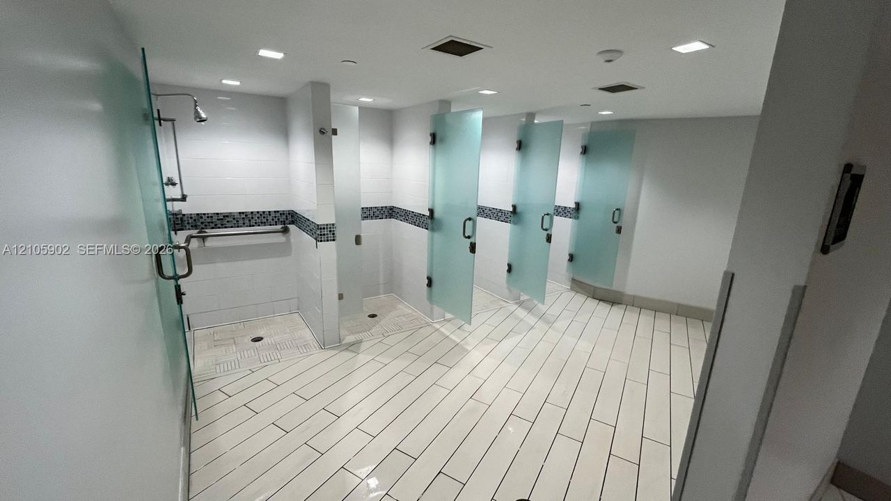Multiple individual shower stalls featuring frosted glass doors, white tiled flooring, inset decorative wall tiling, and recessed ceiling lighting