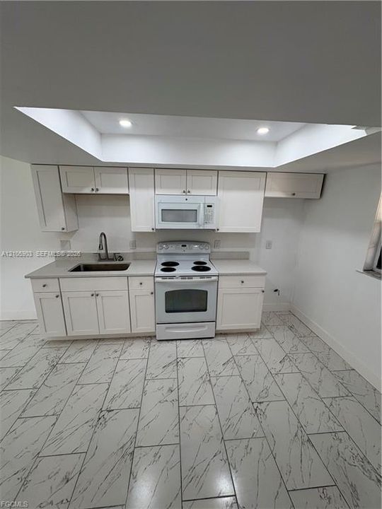 Kitchen featuring white shaker-style cabinetry, solid-surface countertops, a stainless steel sink with a high-arch faucet, a built-in microwave, and patterned tile flooring under a recessed tray ceiling