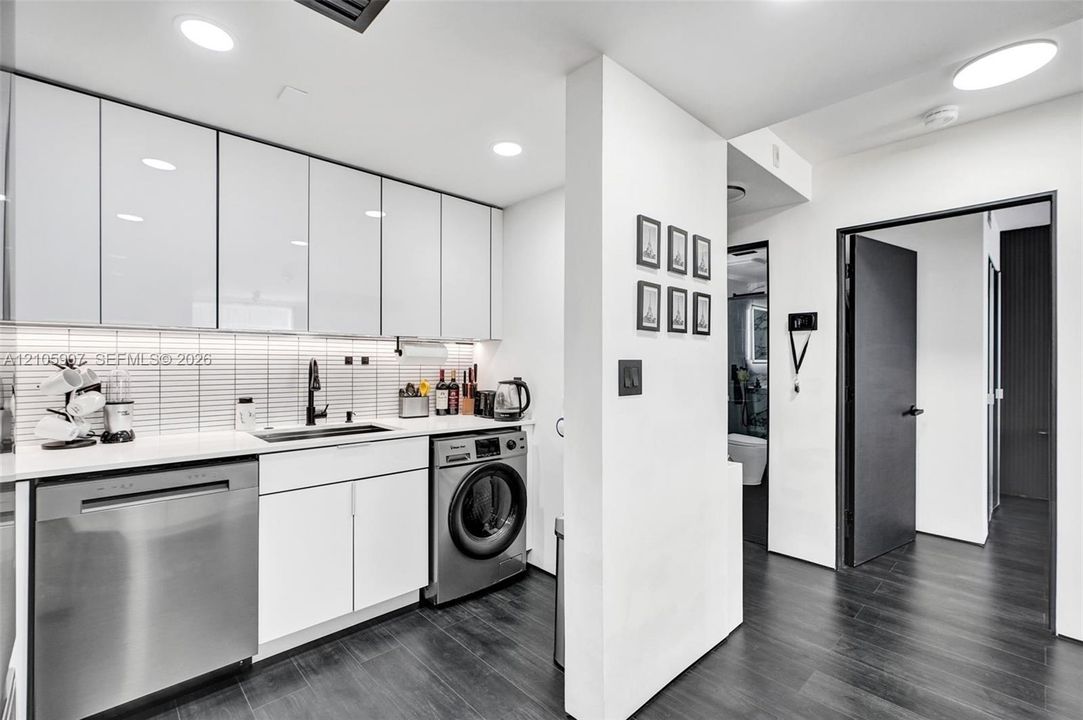 Glossy white cabinetry, linear tile backsplash, stainless steel dishwasher, integrated laundry appliance, and dark-toned flooring