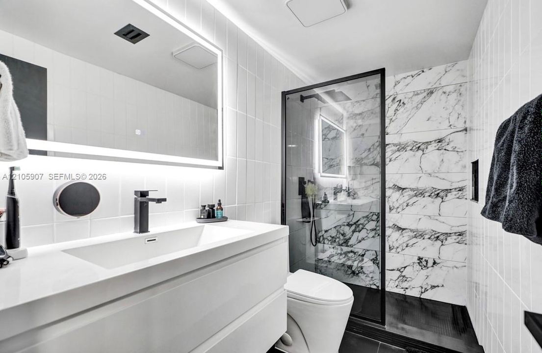 Modern bathroom featuring a floating vanity with an integrated rectangular sink, wall-mounted matte black faucet, illuminated vanity mirror, and a walk-in shower with stone-patterned wall tile and black metal framing