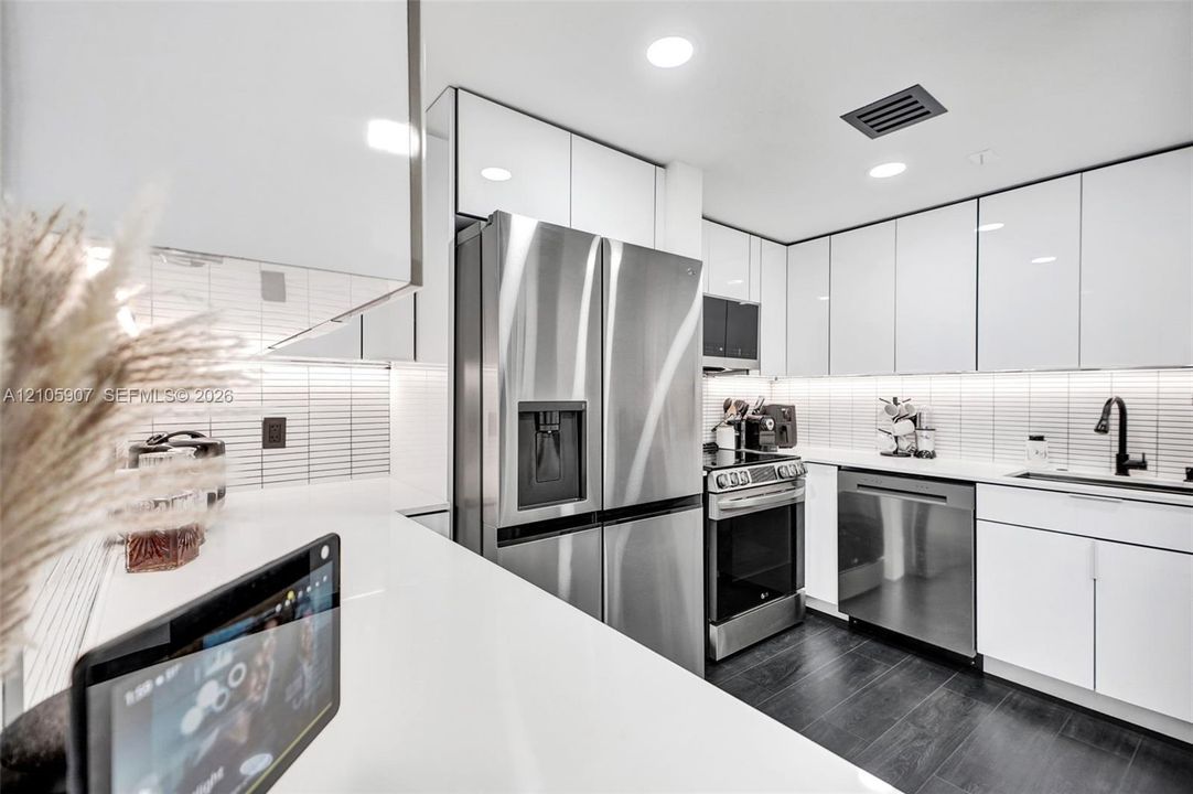 Kitchen featuring high-gloss white cabinetry, stainless steel appliances, white solid-surface countertops, linear tile backsplash, and dark wood-finish flooring
