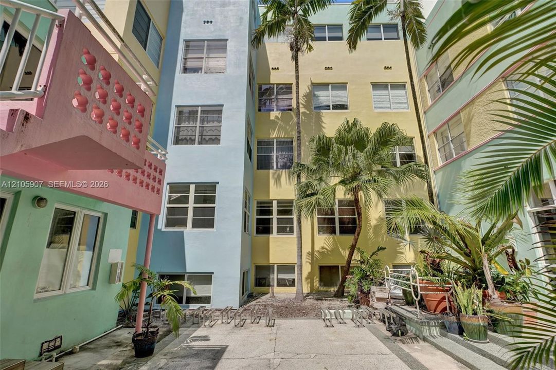 Multi-story pastel exterior featuring geometric concrete breeze block detailing, horizontal casement windows, and a paved courtyard with mature palms