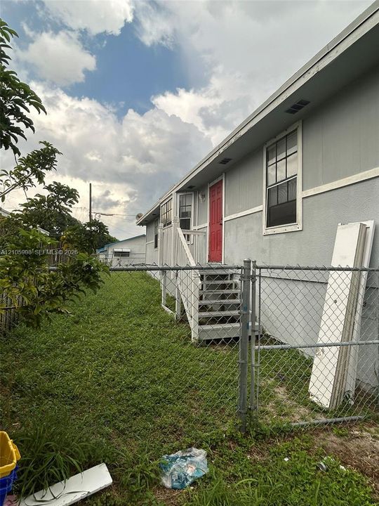 Exterior elevation featuring light-toned siding, white-trimmed windows, a red entry door, and an attached staircase with a white railing system, enclosed by chain-link fencing