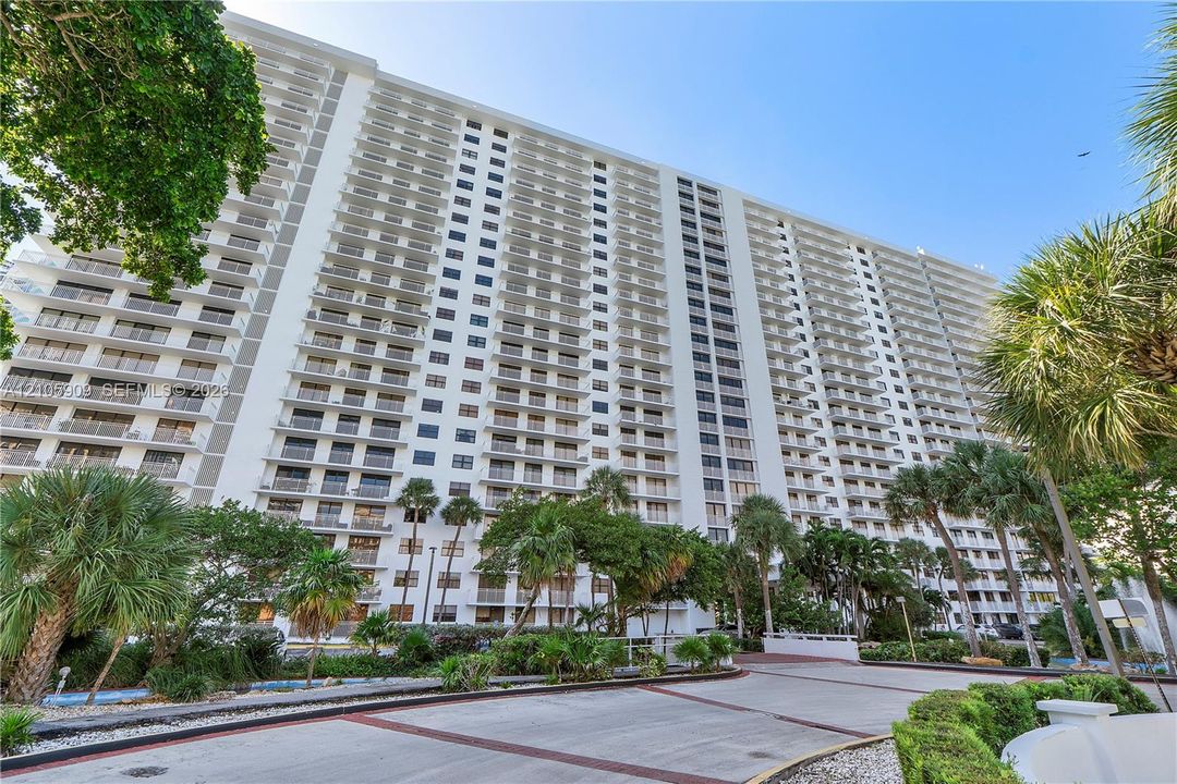High-rise residential exterior featuring expansive balconies and concrete driveway with decorative brick accents, surrounded by mature palms and landscaping