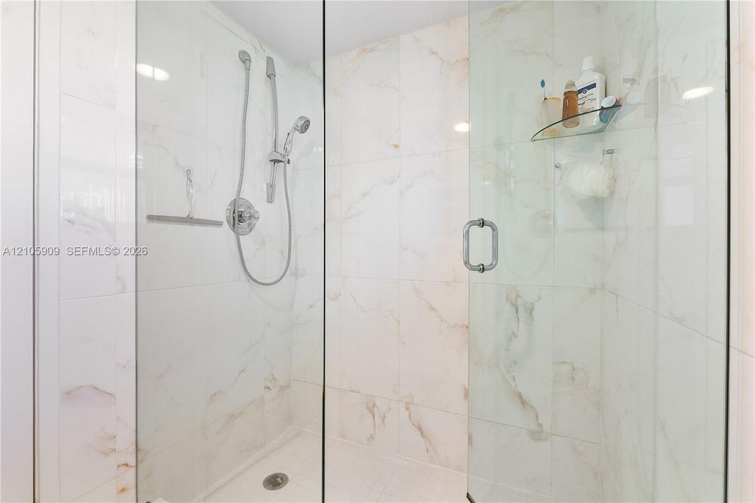 Walk-in shower featuring large-format stone-finish tile surround, frameless glass enclosure, adjustable hand-held showerhead, and integrated glass corner shelving