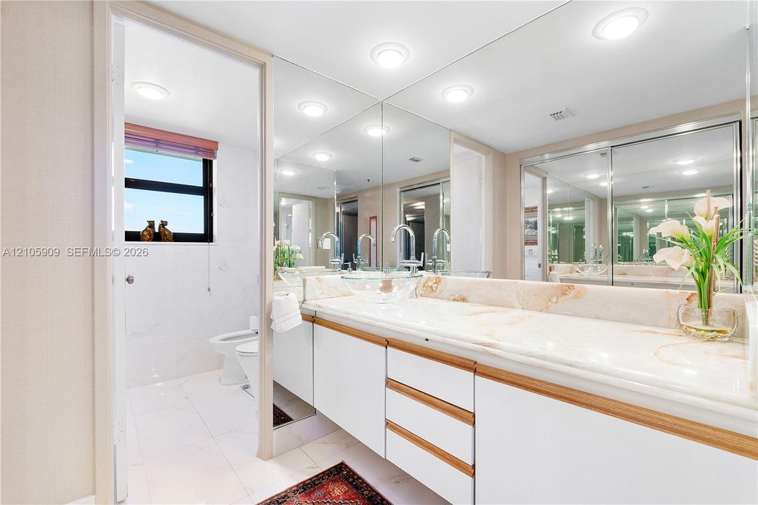 Full-width vanity featuring white cabinetry with wood-tone accents, stone-surface countertop, recessed ceiling lighting, and expansive wall-to-wall mirror