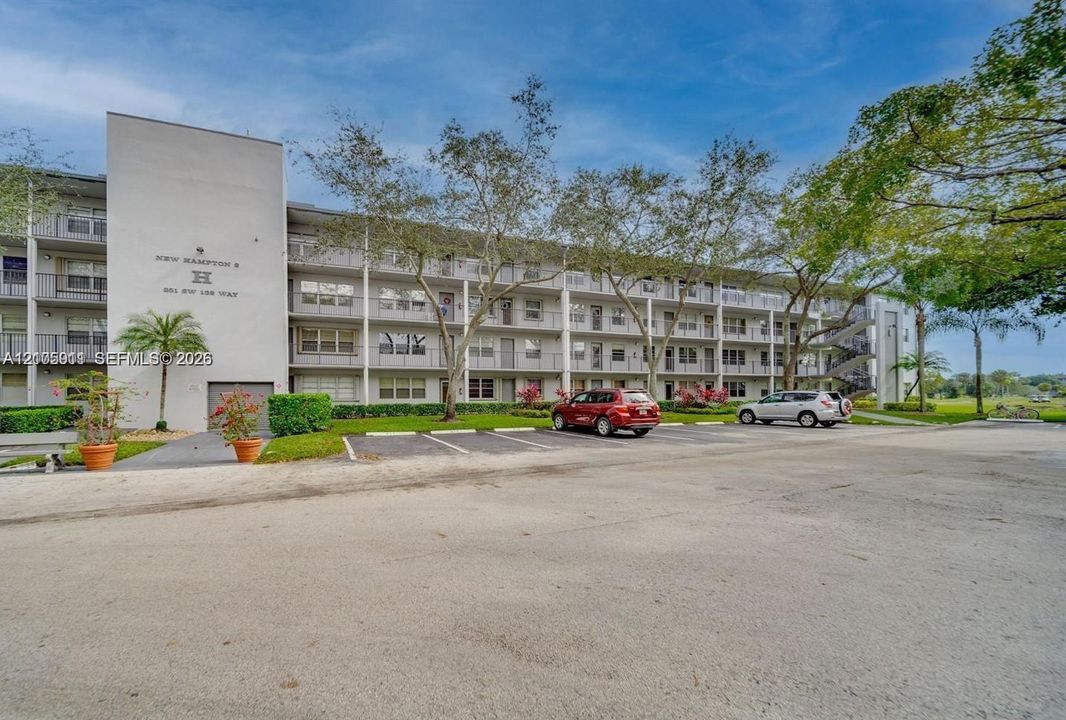 Multi-story residential building exterior featuring concrete facade, private balconies, paved surface parking, and landscaped grounds with mature trees