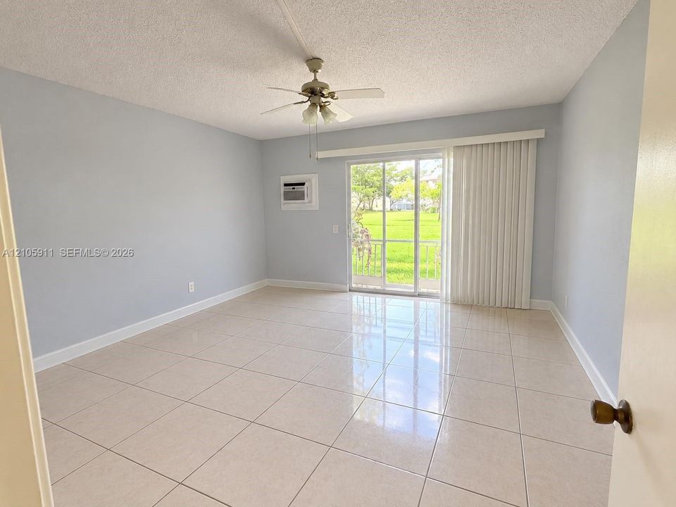 Interior space featuring neutral wall tones, large-format tile flooring, a ceiling fan with integrated light fixture, a wall-mounted air conditioning unit, and sliding glass doors with vertical blinds