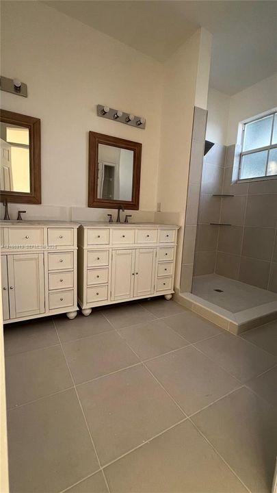 Double vanity with white cabinetry and countertop, two framed mirrors, wall-mounted light fixtures, and a walk-in shower with tile surround