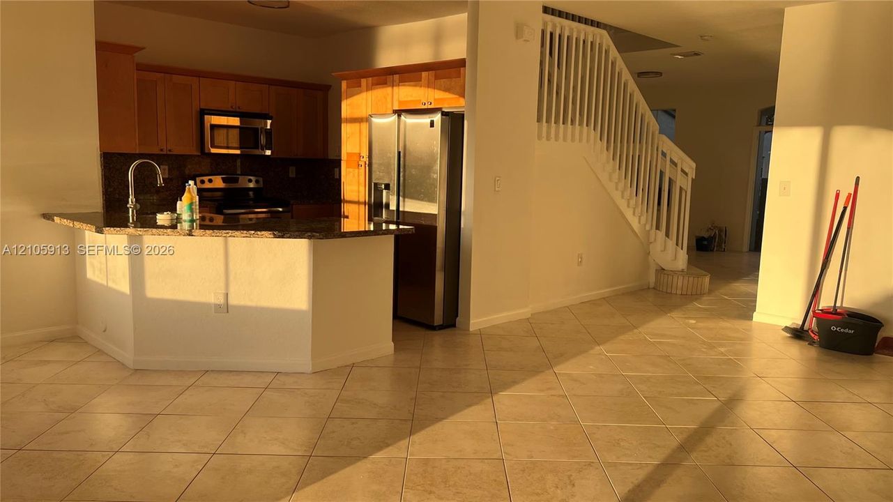 Kitchen featuring wood cabinetry, stone countertops, stainless steel appliances, ceramic tile flooring, and a staircase with a white railing