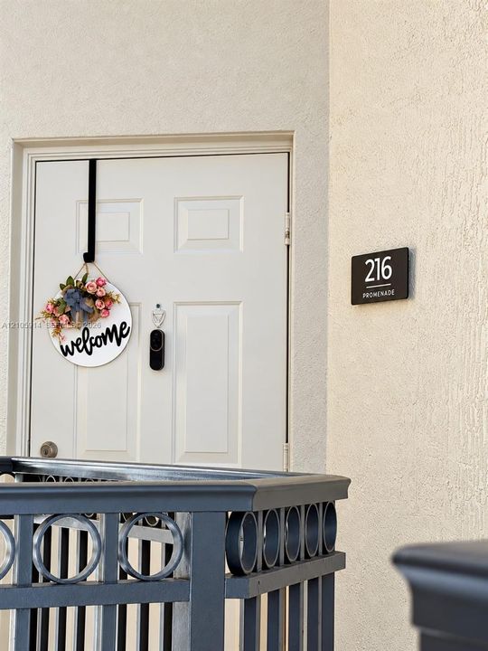 White paneled entry door with integrated peephole and smart doorbell camera, complemented by a matte finish metal railing featuring circular decorative detailing and a mounted address plaque