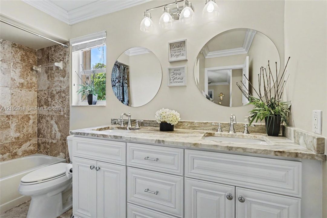 Bathroom featuring a dual-sink vanity with stone countertop, white shaker-style cabinetry, chrome fixtures, dual round wall mirrors, and tiled shower surround