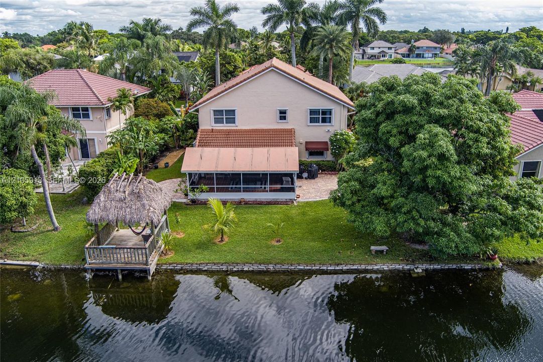 Waterfront two-story residence featuring a screened-in rear lanai, thatched-roof gazebo, paved patio area, and mature landscaping