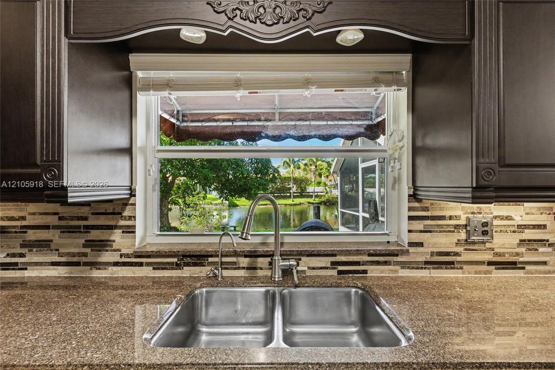Dark wood-finish cabinetry with decorative molding, stone-finish countertops, dual-basin stainless steel sink with high-arc faucet, and multi-tone glass tile backsplash