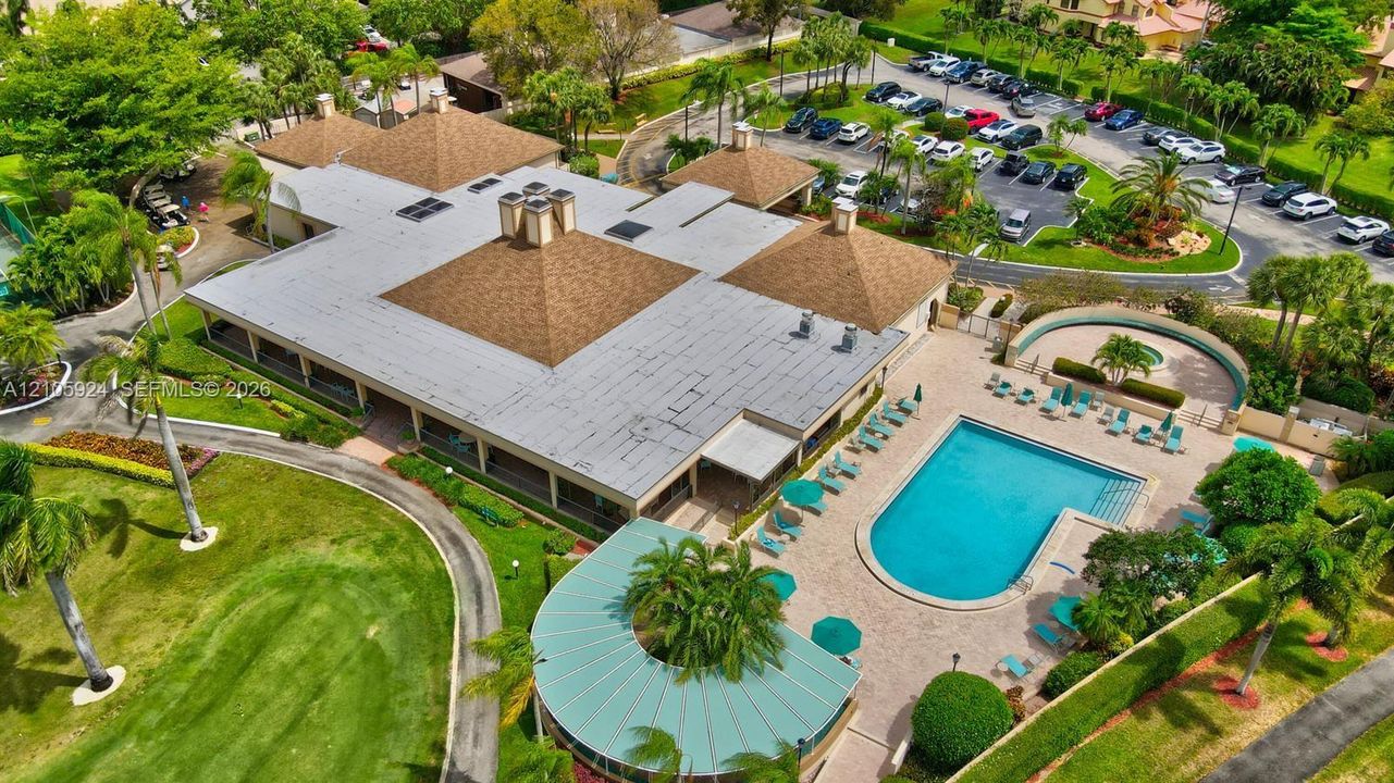 Aerial perspective of a multi-section structure featuring a shingled hipped roof, an expansive paved pool deck, a rectangular swimming pool, and manicured landscaping with palm trees
