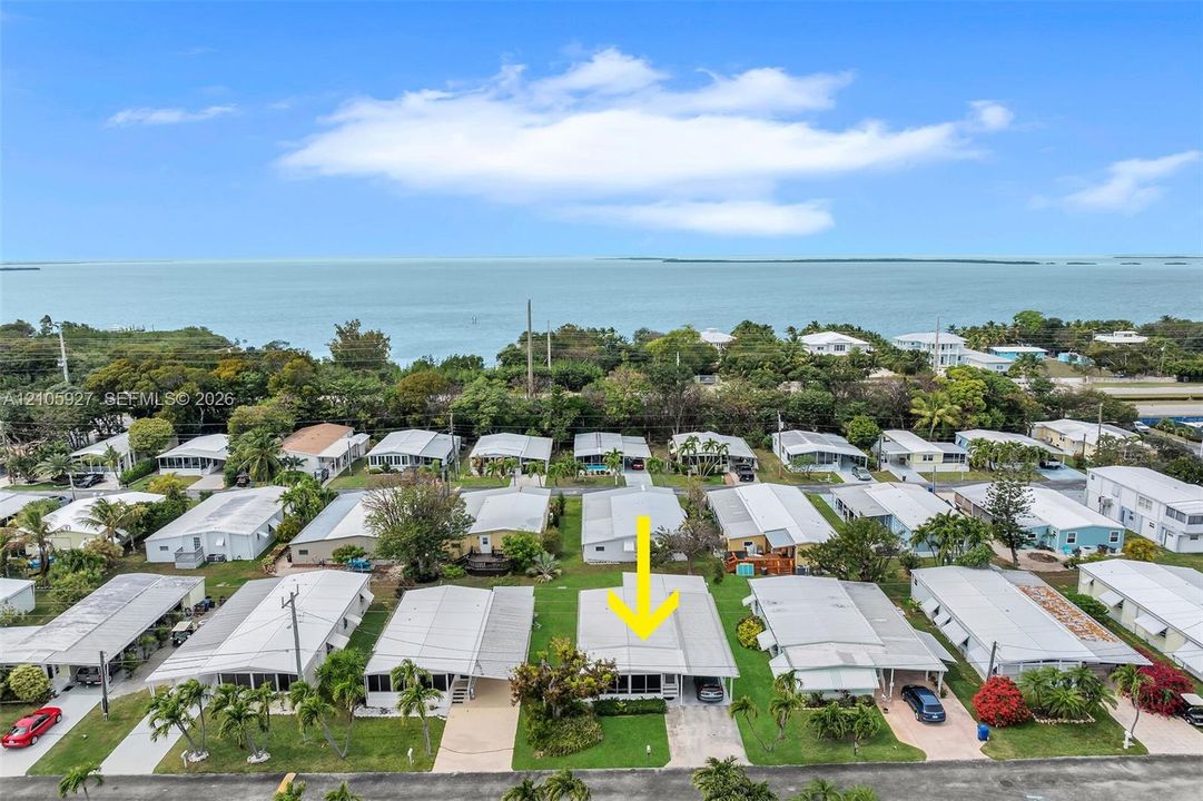 Aerial perspective featuring a single-story residence with a metal roof, attached carport, and a screened-in porch area, situated in a coastal setting with proximity to the shoreline