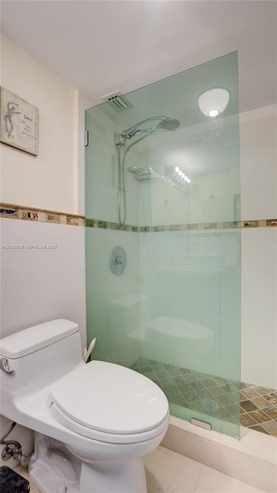 Walk-in shower featuring a frameless glass partition, wall-mounted rainfall showerhead, decorative tile border, and mosaic stone-finish floor