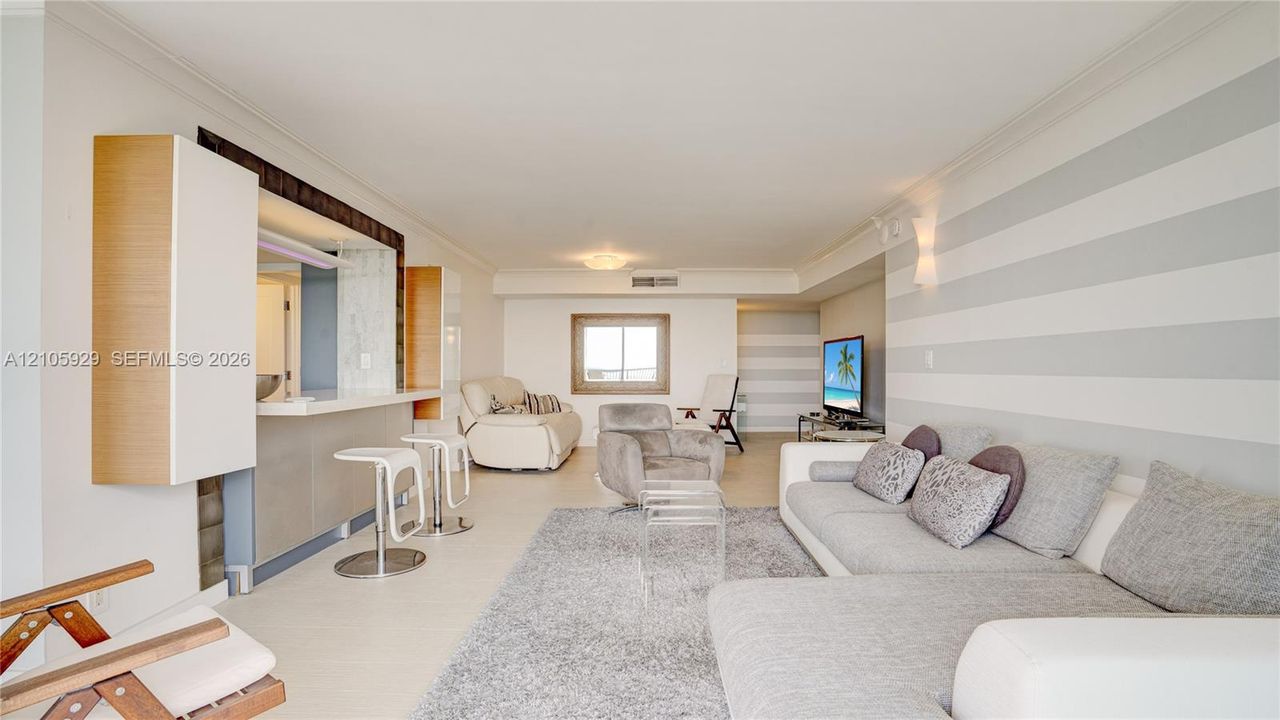Living area featuring a service pass-through with a breakfast bar, neutral-tone striped wall treatments, crown molding, and large-format tile flooring