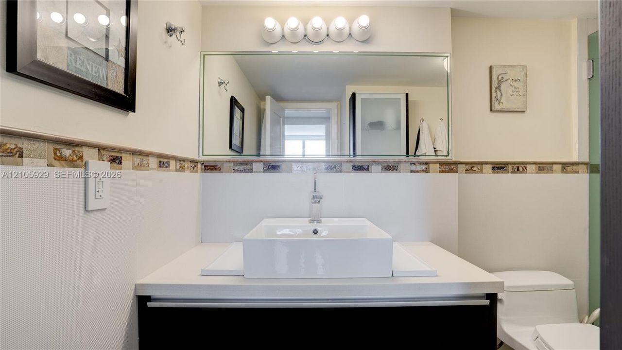 Vanity featuring a rectangular vessel sink, solid-surface countertop, decorative tile backsplash, wall-mounted mirror, and multi-bulb light fixture