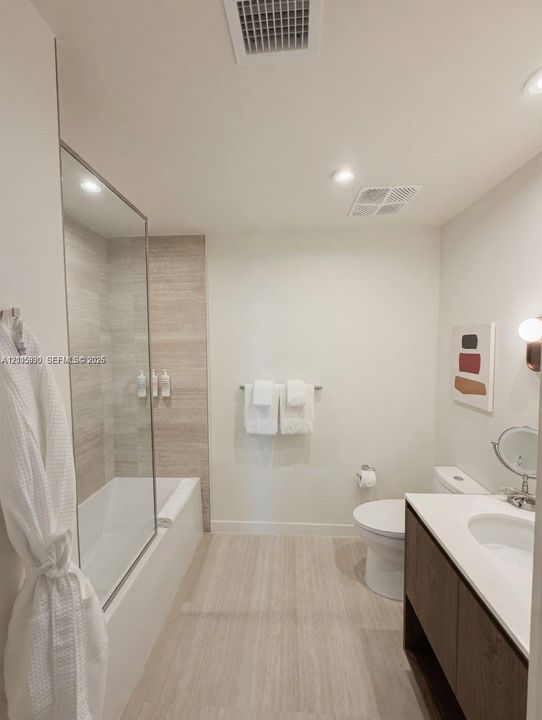 Bathroom featuring a vanity with dark wood-finish cabinetry, a white countertop with an integrated basin, a soaking tub with a glass panel enclosure, and horizontal stone-patterned wall tiling