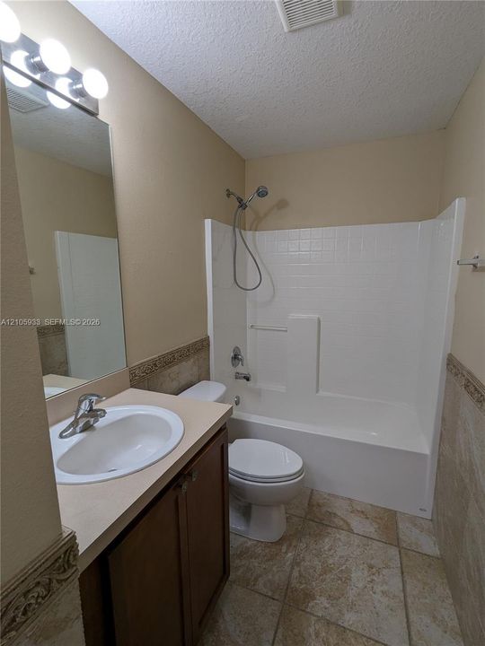 Bathroom featuring a vanity with an integrated sink, stone-tile flooring, wall-mounted mirror with multi-bulb lighting, and a white acrylic shower-tub combo