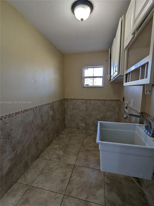 Utility area featuring large-format ceramic tile flooring, partial-height ceramic wall tiling with mosaic accent border, white upper cabinetry, and a deep basin utility sink with dual-handle faucet