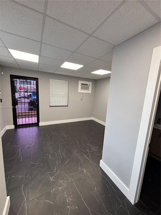 Dark large-format tile flooring with veining detail, recessed ceiling lighting panels, wall-mounted air conditioning unit, and metal security storm door