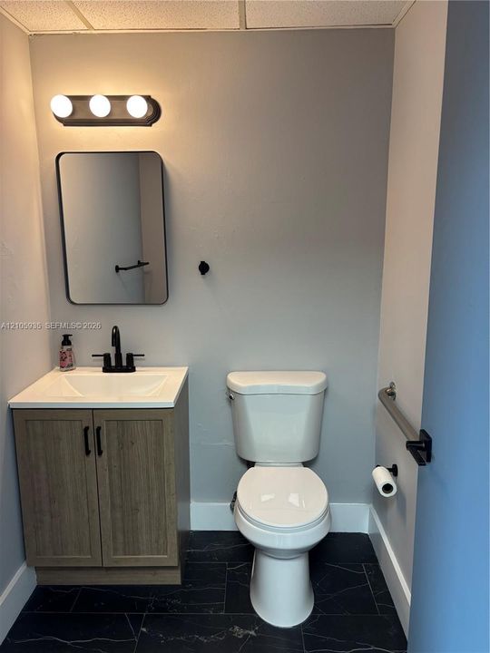 Bathroom featuring a vanity with wood-finish cabinetry, a white integrated sink, a three-bulb wall-mounted light fixture, a rectangular mirror with black framing, and matte black hardware