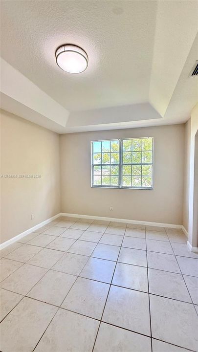 Tray ceiling with central flush-mount lighting, large grid-pane window, and neutral ceramic tile flooring