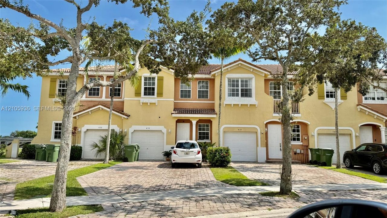 Multi-story townhome exterior featuring a stucco facade, tile roof, arched garage entries, and a decorative brick-paver driveway